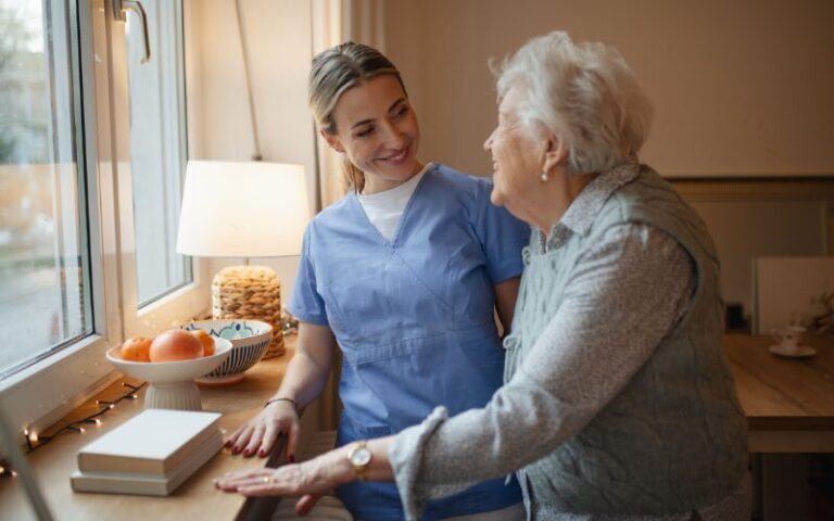 Healthcare worker smiling and interacting with elderly woman in a warm, inviting senior living environment, emphasizing compassionate care and support for aging individuals.